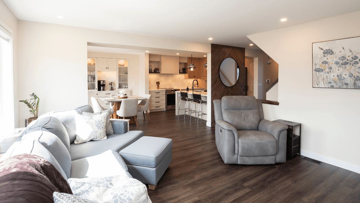A view of a modern open-concept main floor. The living room, featuring a gray sectional sofa and a recliner, flows into a dining area with a round table and a kitchen. The kitchen renovation includes light-colored cabinets, a dark island, and a decorative wall with a round mirror.