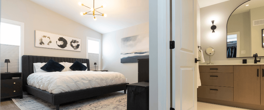 A renovated master bedroom with a large bed and a glimpse into a modern ensuite bathroom with a dark vanity and a large mirror.