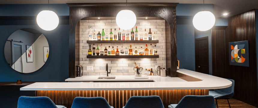 Renovating your forever home: A custom-built basement bar renovation with a curved white countertop, blue velvet bar stools, a white brick backsplash, and dark wood trim.
